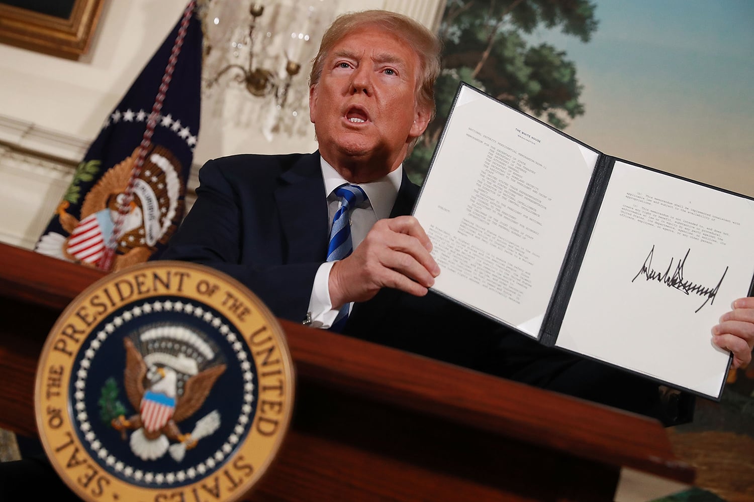 A man in a dark suit and blue striped tie sits at a desk, holding up an open folder. The folder contains a white document with a prominent, jagged black signature at the bottom. A large gold seal of the President of the United States is visible in the lower-left foreground.