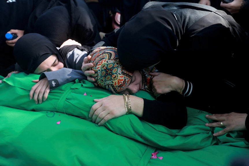 Three women in head coverings lean over the covered remains of a person, wrapped in green cloth, during a funeral.