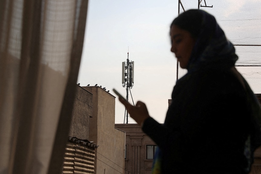 A woman in a headscarf looks at her phone while standing in an apartment in a city.