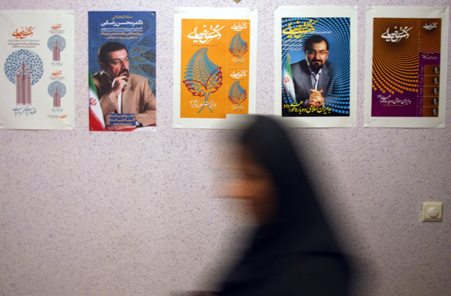 Five political posters are mounted in a row on a light-colored, textured wall. The posters feature portraits of a man and various graphic designs in blue, orange, and purple. In the foreground, the blurry silhouette of a person in a dark headscarf walks past, creating a sense of motion.