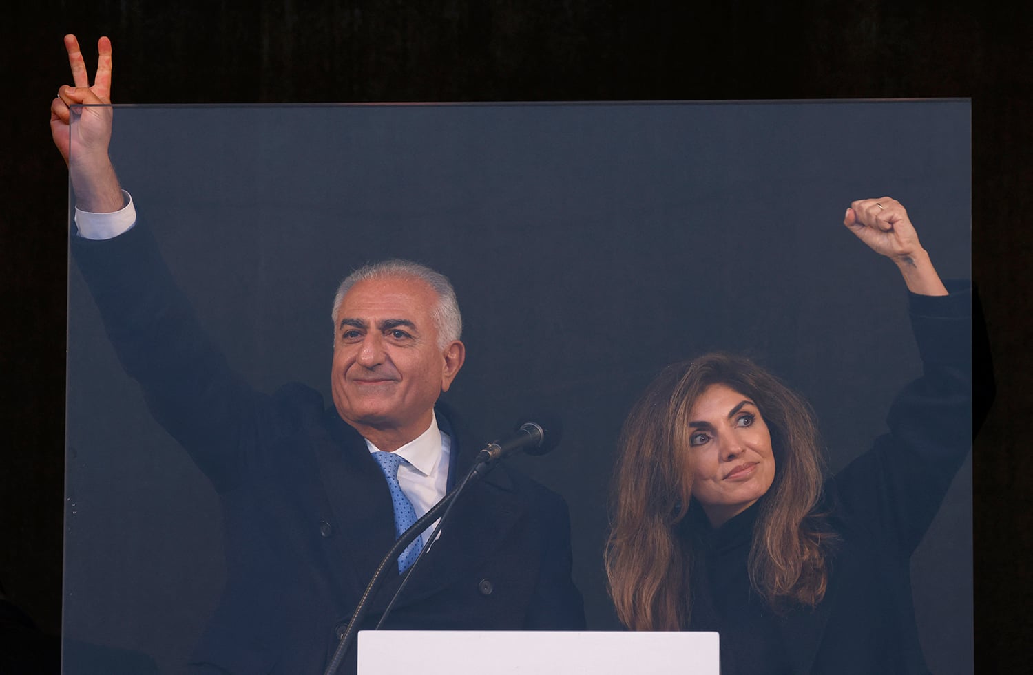 A man and a woman stand side-by-side behind a clear protective barrier. The man on the left wears a dark coat, white shirt, and blue tie, raising his right hand in a peace sign. The woman on the right wears a dark top and raises her left fist in the air.