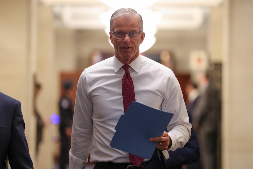 Thune walks down a hallway holding a blue manila folder.