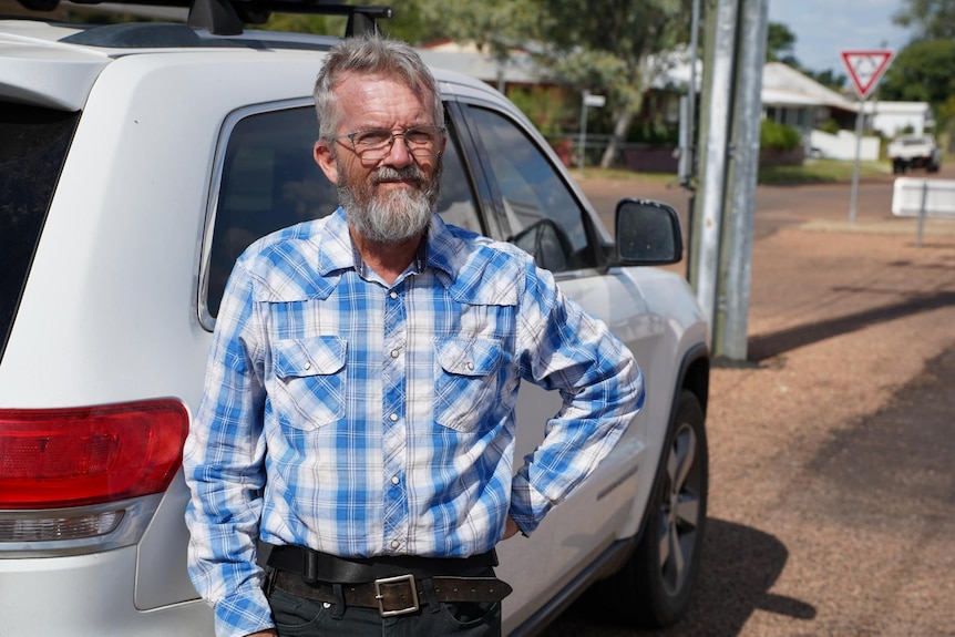 A man in glasses and a blue and white checked shirt smiles near his white car.