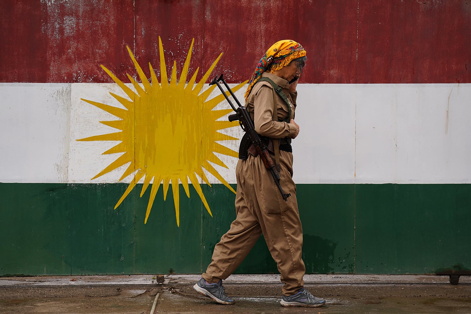 A side profile of a person in tan military-style clothing and a colorful yellow headscarf walking past a large wall painted with the Kurdish flag—featuring horizontal bands of red, white, and green with a large yellow sun in the center. A rifle is slung across the person's back.