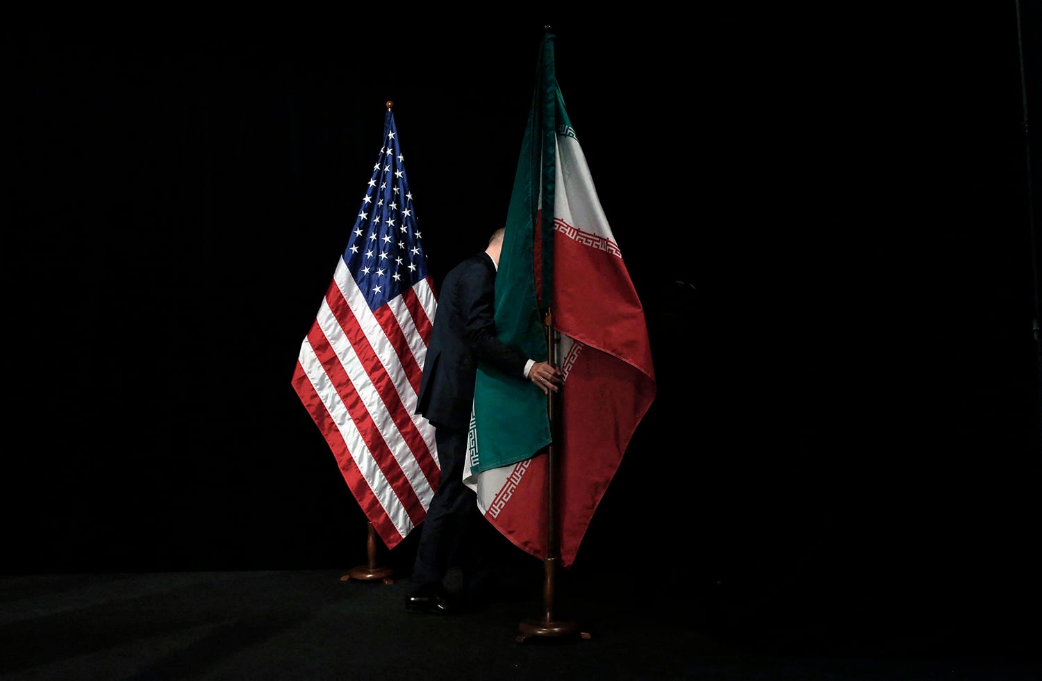 The American and Iranian flags stand side-by-side on wooden poles against a black background. A person in a dark suit is partially visible behind the Iranian flag, reaching out to adjust or hold the flagpole, their face obscured by the fall of the flag.