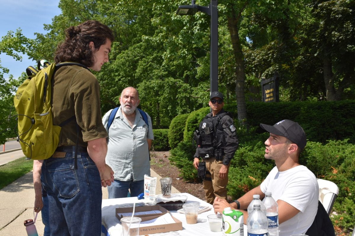 A person seated behind a table talks to a student. A security guard stands in the background.
