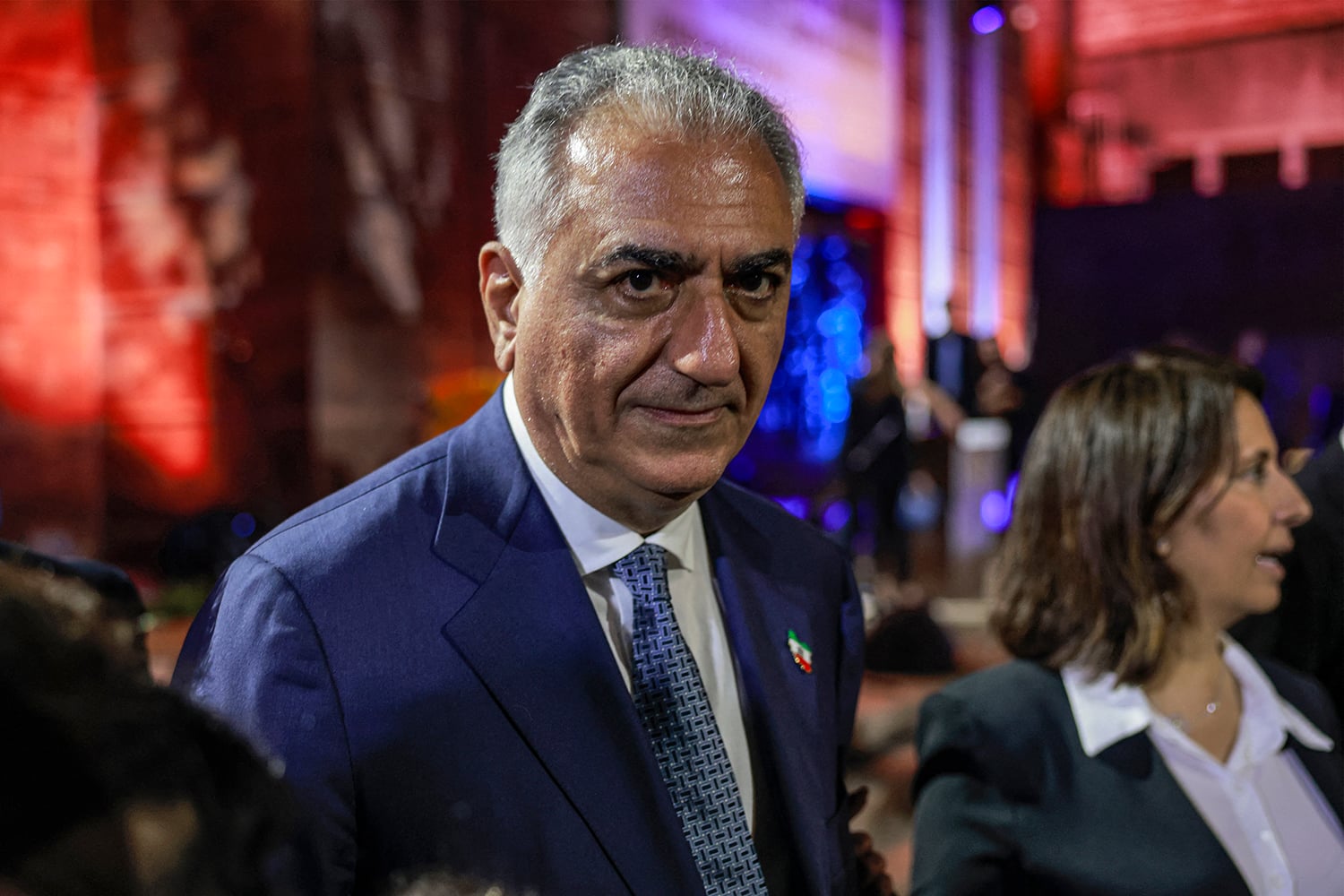 A close-up, slightly low-angle photograph of a man in a dark blue suit, white collared shirt, and patterned tie. He has short graying hair and is looking directly toward the camera with a serious expression. The background is dimly lit with red and blue ambient lights and other indistinct figures.