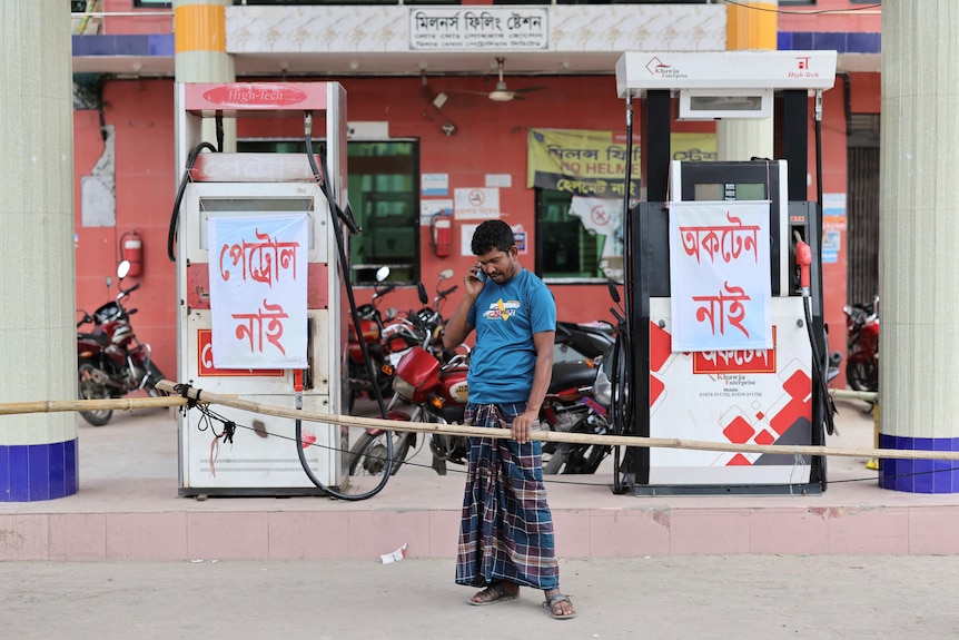 A man stands and talks on the phone at a petrol station, where signs indictate a fuel shortage.