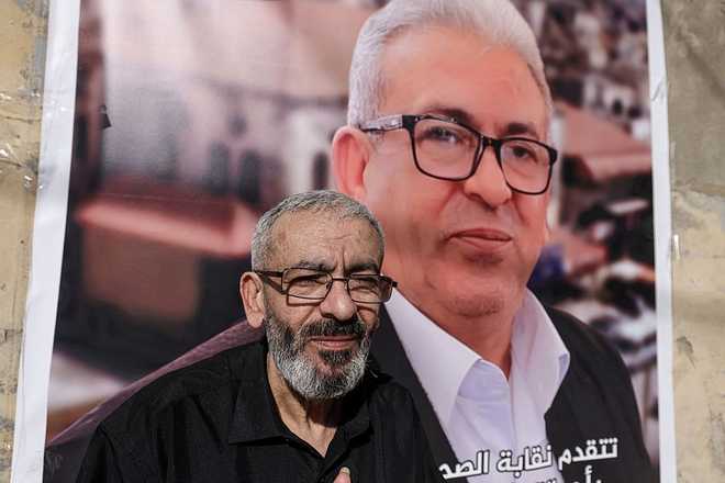 PALESTINIAN-ISRAEL-CONFLICT-MEDIA-PRISONER Palestinian journalist Ali al-Samoudi stands in front of a large image of himself taken before he was detained by the Israeli army and held for a year, at his home in the northern city of Jenin, in the Israeli-occupied Palestinian West Bank on May 1, 2026. Sixty-year-old Al-Samoudi was released following one-year in detention in the Israeli Naqab Prison, located in southern Israel. He was released and checked at the Jenin Hospital where he said, "I was 120 kilogrammes; my weight now is barely 60 kilogrammes". Israel has jailed an increasing number of journalists since 2023, the year of Hamas's unprecedented October 7 attack on Israel, which responded with a devastating war in the Gaza Strip. (Photo by JAAFAR ASHTIYEH / AFP via Getty Images)