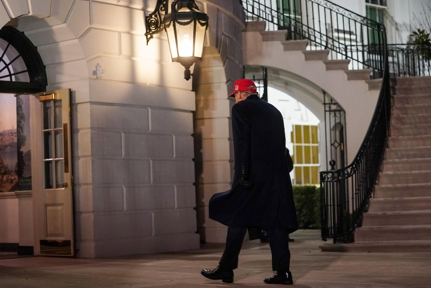 Wearing a black trench coat and red baseball cap, Trump walks inside the White House, which is lit up at night