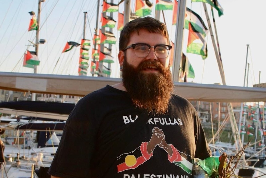A man with a beard and glasses, standing on a boat with Palestinian flags in the background.