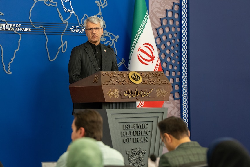 A man with grey hair stands behind a lectern that says Islamic Republic of Iran