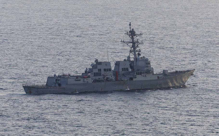 A military destroyer, seen at a distance and from a high angle, sails in open ocean with the blurry horizon line in the background.
