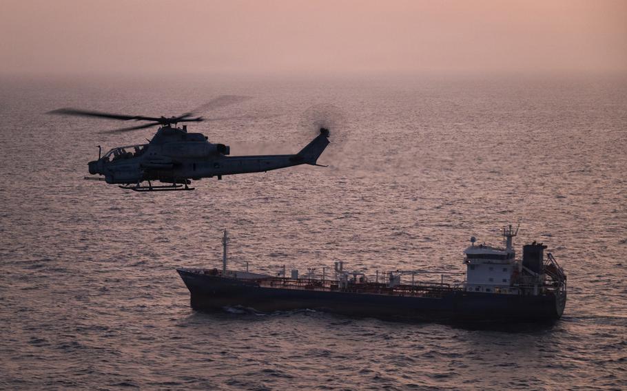 A military helicopter flies above a ship sailing in open ocean, with the setting sun and horizon line in the background.