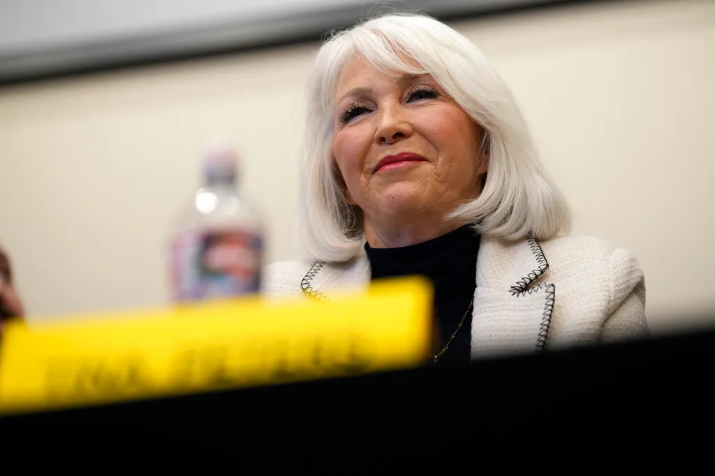 Tina Peters, who has white hair, is seated, wearing a white jacket and black top, with a water bottle and a yellow name tag in front of her.