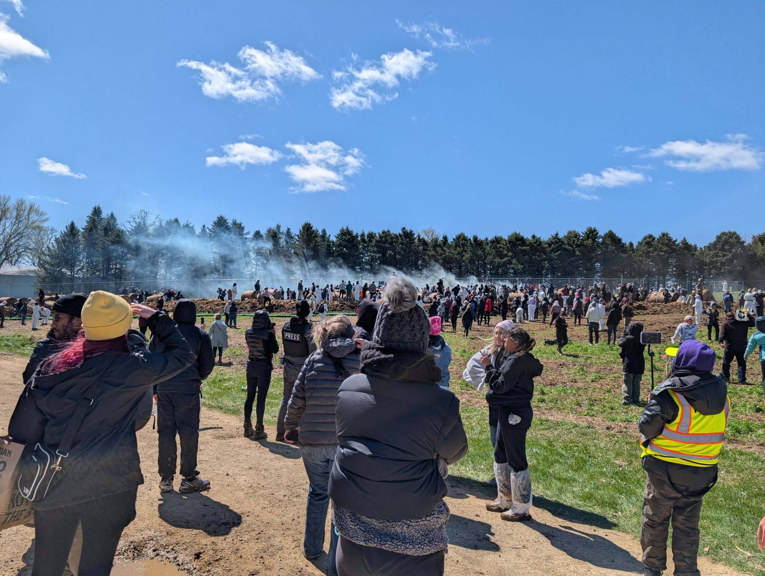 A large crowd gathers outdoors on a dirt path, with some people standing and others facing smoke in the distance under a partly cloudy sky.