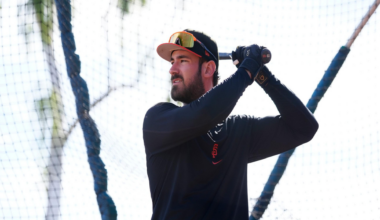 A man wearing a black long-sleeve shirt, black gloves, and a baseball cap holds a bat, preparing to swing inside a batting cage.