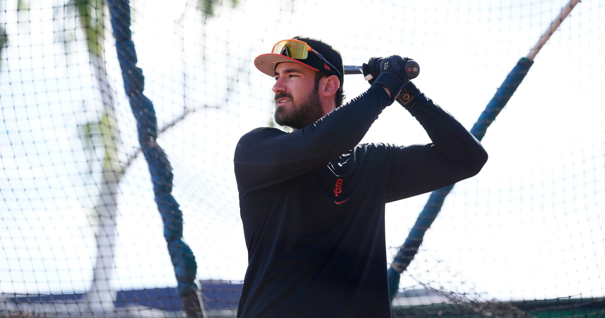 A man wearing a black long-sleeve shirt, black gloves, and a baseball cap holds a bat, preparing to swing inside a batting cage.