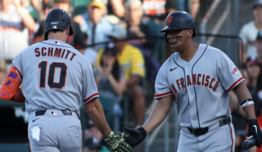 Two San Francisco Giants players in gray uniforms share a mid-game handshake, one holding a bat and the other wearing an orange arm sleeve.