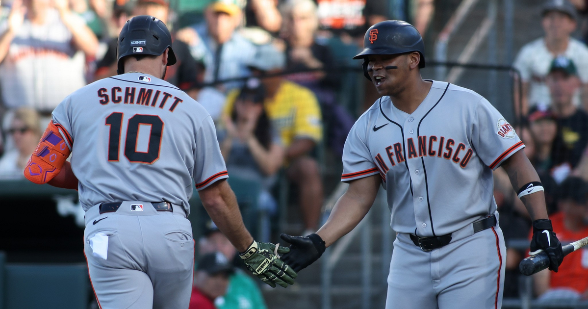Two San Francisco Giants players in gray uniforms share a mid-game handshake, one holding a bat and the other wearing an orange arm sleeve.