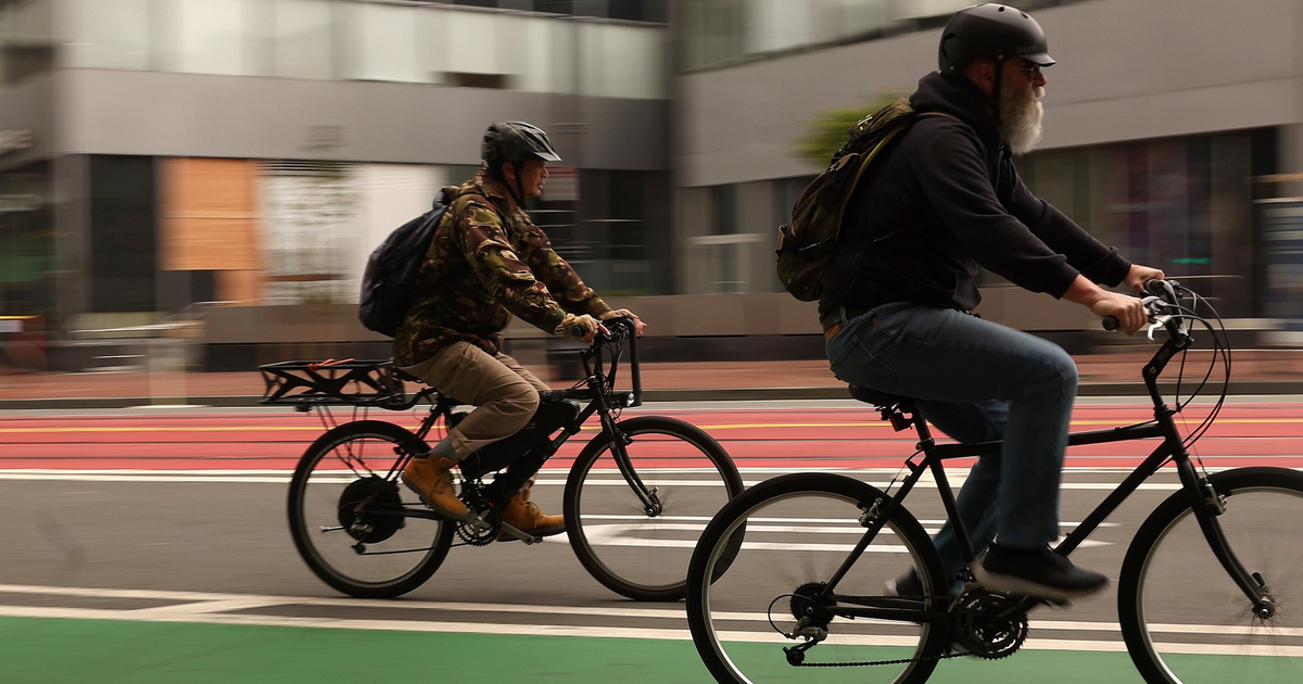 Two men wearing helmets ride bicycles on a green bike lane in an urban area with blurred buildings in the background.