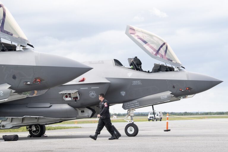 The nose cones and open hatches of two F-35 fighter jets are visible in this shot. The aircraft are parked on asphalt and there is a ground-crew member in coveralls and sunglasses walking past them, pulling a toolkit on wheels.