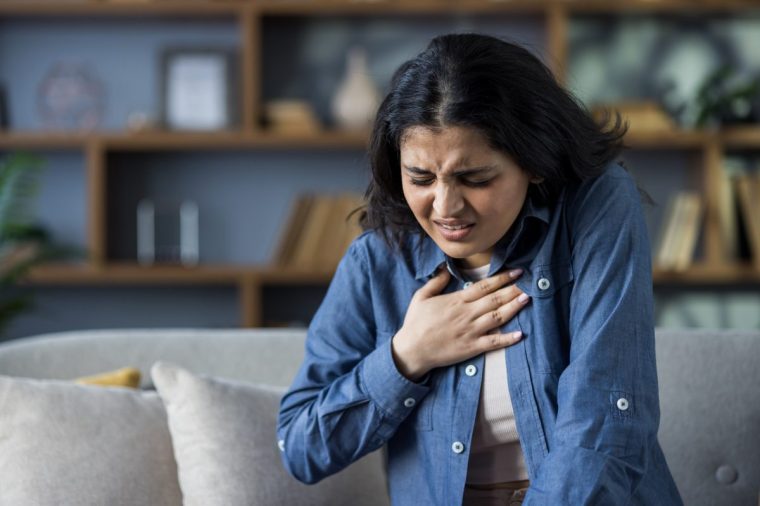 A young Indian woman sits on a couch at home, holding her hand over her heart, grimacing as she feels severe pain in her chest - stock photo
