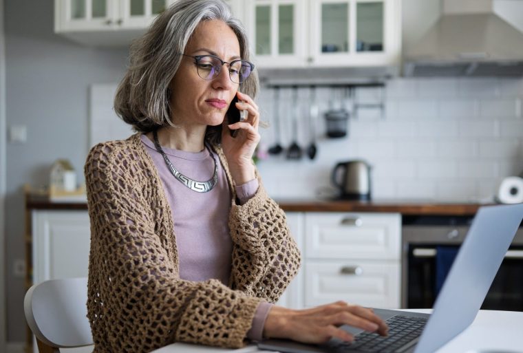 Mid adult businesswoman using phone and laptop for work from home. - stock photo