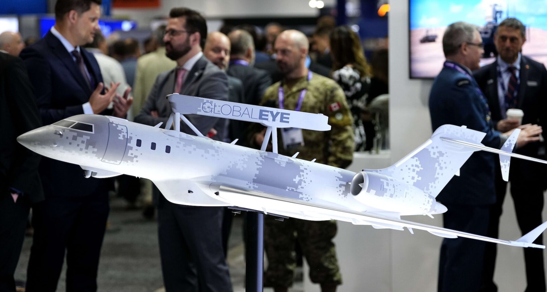A closeup of a scale model of a jet covered in pixellated camouflage, with sensor equipment attached to the top of its fuselage. There are civilians and uniformed military personnel milling in the background.