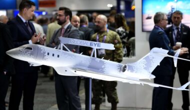 A closeup of a scale model of a jet covered in pixellated camouflage, with sensor equipment attached to the top of its fuselage. There are civilians and uniformed military personnel milling in the background.