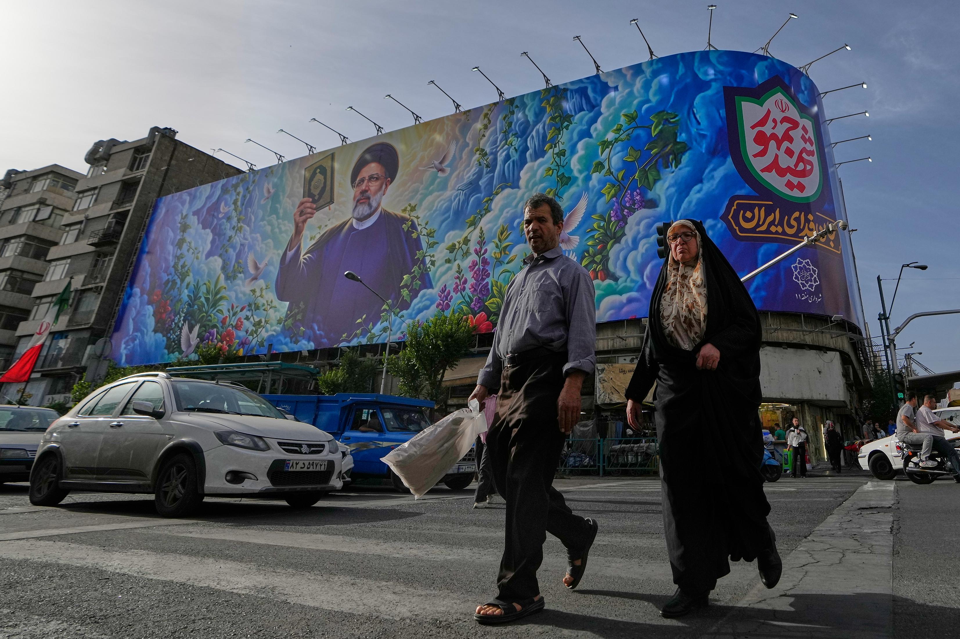 People cross an intersection in front of a billboard of the late Iranian President Ebrahim Raisi.