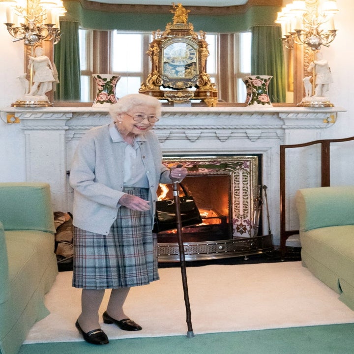 An elderly woman stands in a cozy room with a fireplace, wearing a plaid skirt and cardigan, holding a cane