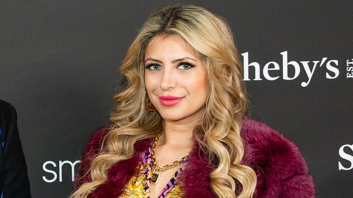 Jasveen Sangha wearing a purple fur top posing at a step and repeat backdrop