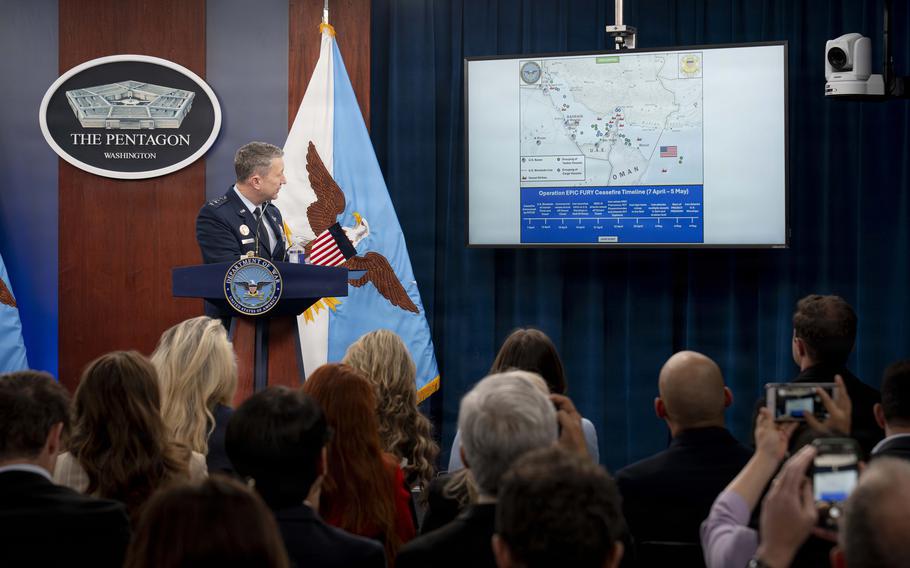A U.S. Air Force general in service dress stands at a Pentagon podium beside a screen displaying a map and ceasefire timeline, addressing a crowd of reporters holding cameras and phones.