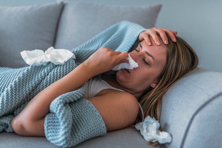 Sick Woman having Flu with Caught Cold. Sneezing into Tissue. Headache. Virus. Sick woman lying in bed with high fever. She is blowing nose. Photo of Young female Lying On Bed covered with a blanket Infected With Allergy holding her head.