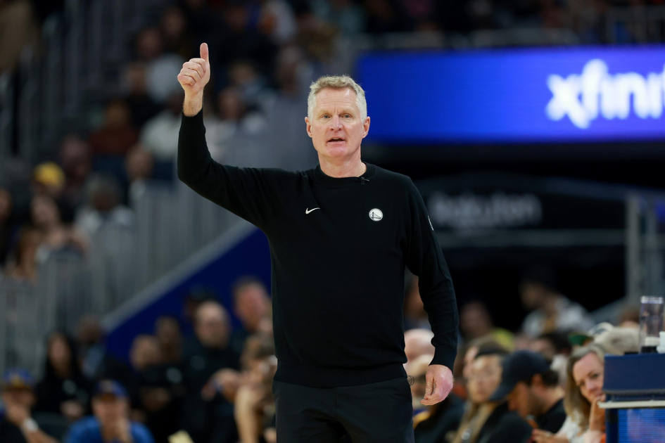 A man in a black long-sleeve shirt with a small logo raises his right hand with a thumbs-up during a crowded event.