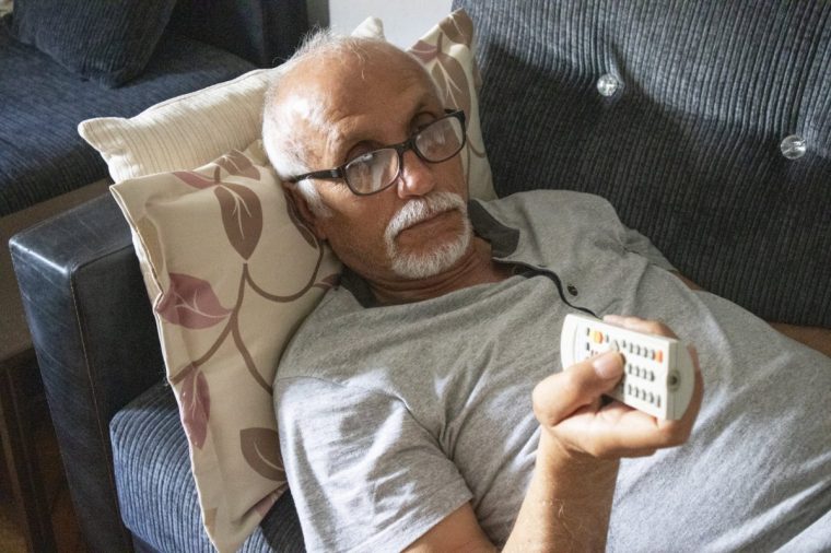 Senior man laying down on the sofa, watching tv and holding the remote control.