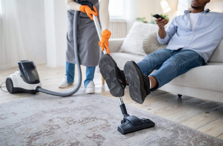Millennial black lady vacuuming floor while her husband watching TV, not helping her with household duties, cropped view. Unrecognizable young couple having problem with domestic chores