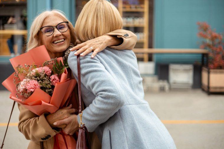 Heartwarming lifestyle image of adult daughter and senior mother sharing a joyful embrace with a bouquet of flowers on a city street, highlighting love, connection, and hugging.