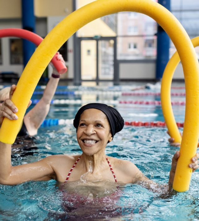 Healthy senior woman doing swim noodle exercises in aerobics pool class. Active senior people doing workout in swimming pool holding swim noodles.