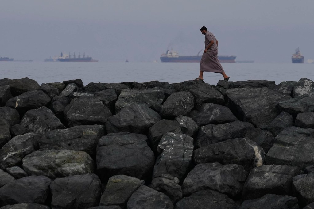 A man walks along a large wall of boulders. Oil tankers and cargo ships are sailing on the sea in the background.