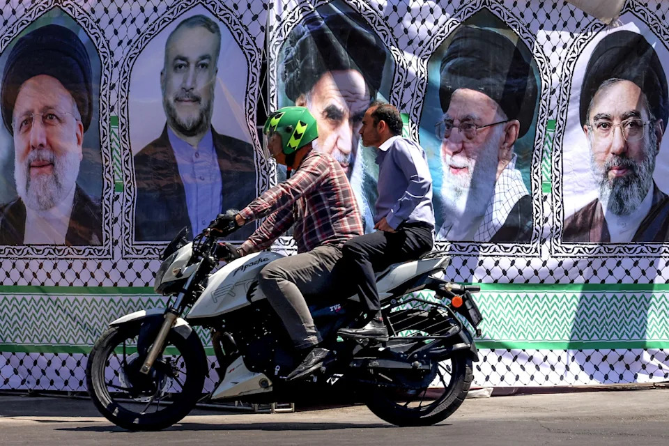 Atta Kenare/AFP via Getty Images - PHOTO: A man rides his motorbike past a banner depicting Iranian leaders in Tehran on May 25, 2026.