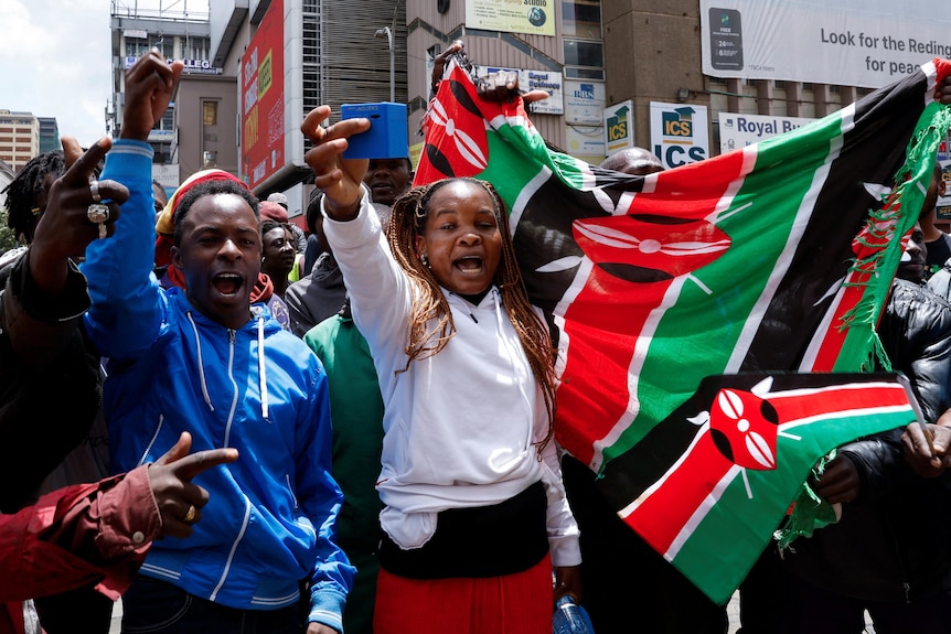 A man and woman, along with a bigger crowd, chant in the street while holding up a Kenyan flag.