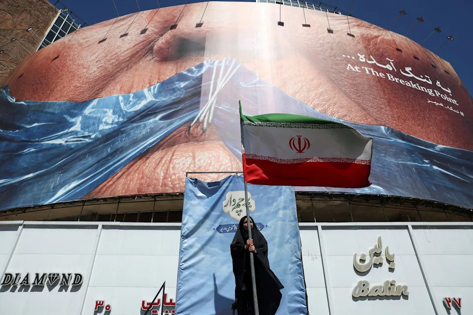 Majid Asgaripour/via Reuters - PHOTO: A woman holds an Iranian flag near an anti-U.S. billboard depicting U.S. President Donald Trump and the Strait of Hormuz, in Tehran, Iran, on May 25, 2026.