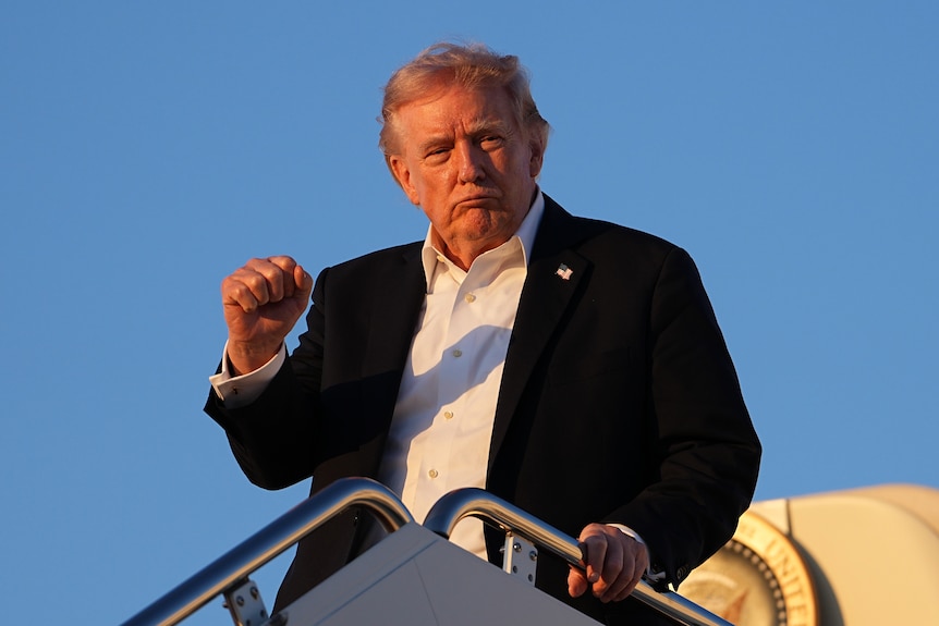 A man in black suit jacket and white shirt, is leaving a plane. He holds his right hand up in a closed-fist.