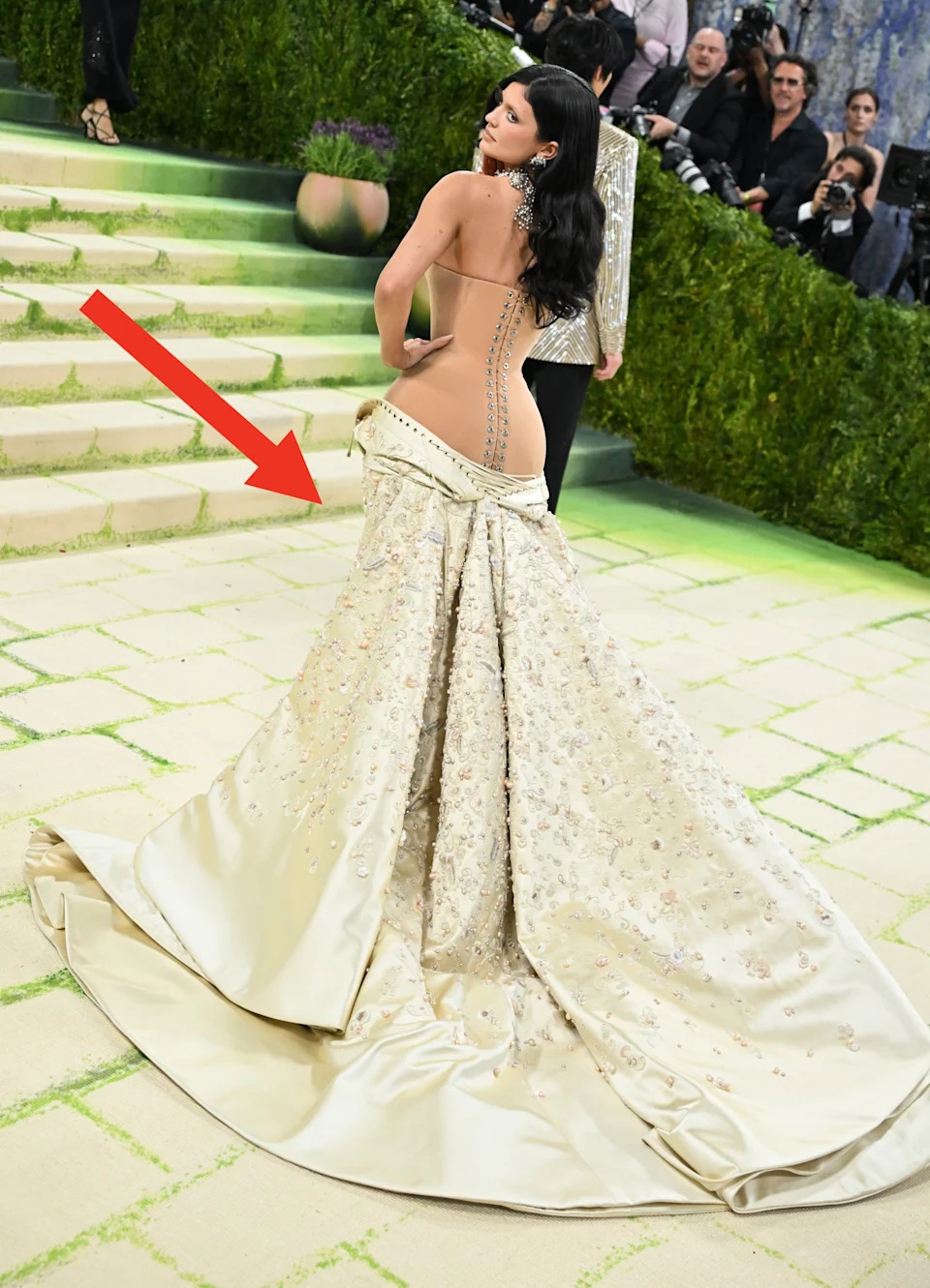 Kylie Jenner in an ornate strapless gown with a detailed train on a carpeted runway, surrounded by photographers