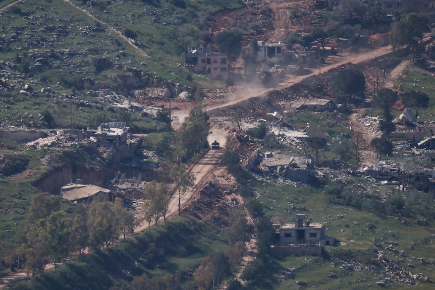 Aerial view of an IDF military vehicle driving down a dirt road past a series of bombed out buildings