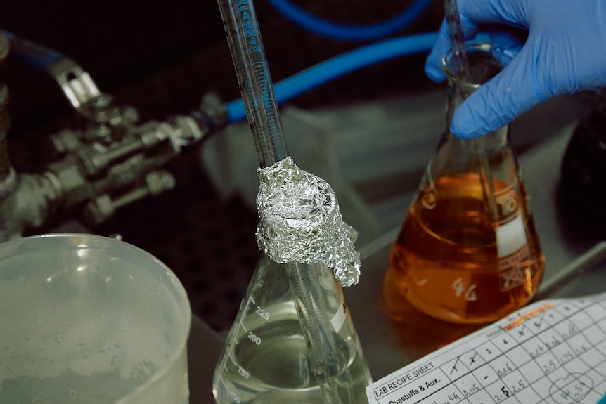 beakers of coloured and clear liquid on a metal work bench both have glass measuring instruments in them one is being picked up