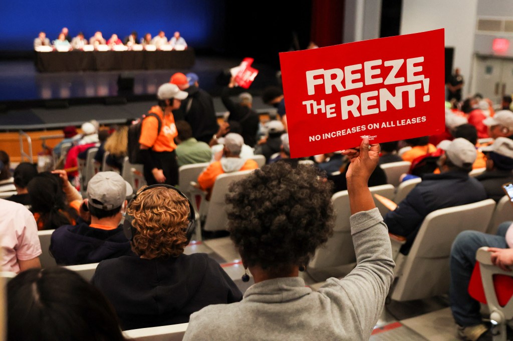 A person holds up a red sign that reads "FREEZE THE RENT! NO INCREASES ON ALL LEASES" at a public hearing.