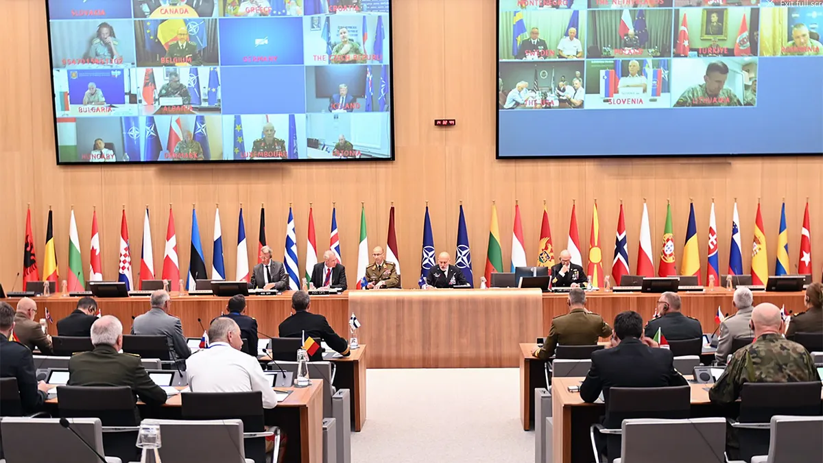 NATO Chiefs of Defense holding a hybrid meeting with screens showing allied leaders joining remotely